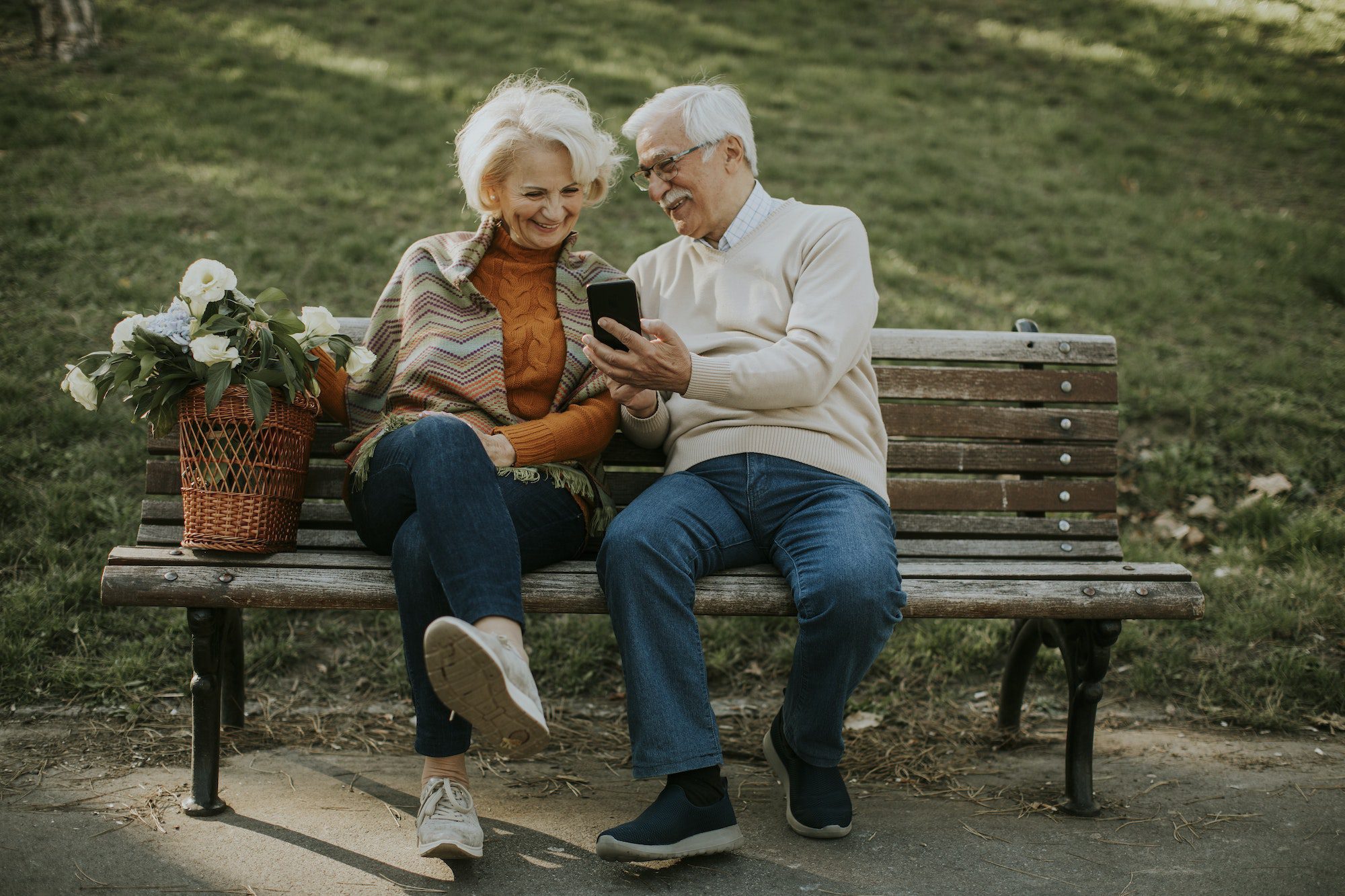 Senior couple sitting on the benchwith basket full of flowers and lookin at mobile phone checking on Medigap Premium increasing